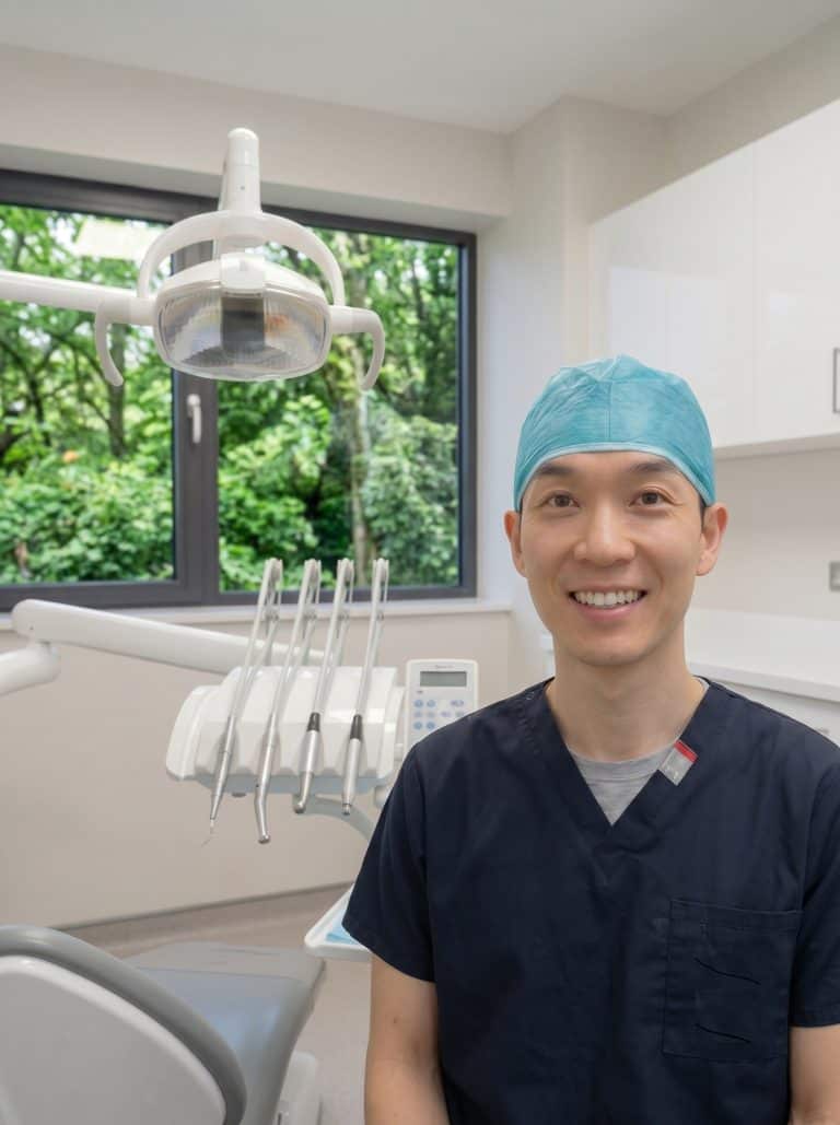 Dentist in navy scrubs and teal cap smiling beside a dental chair in a bright clinic with an overhead light and dental tools nearby.