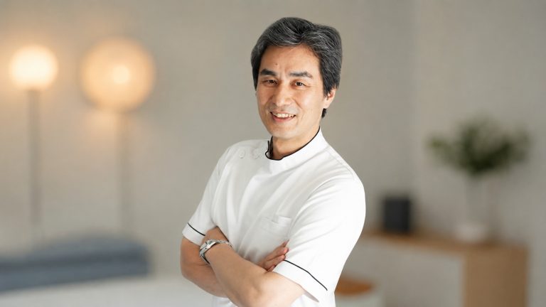 Smiling male healthcare professional in a white coat with arms crossed in a bright, modern clinic setting.